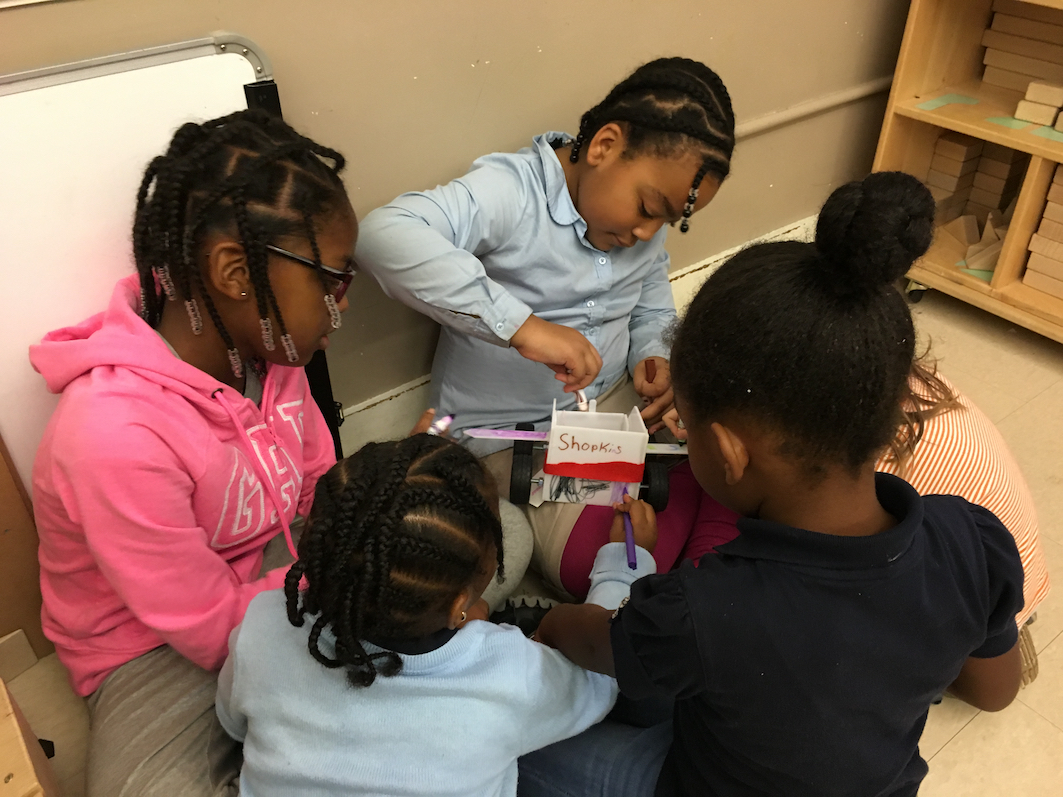 A group of children gather around a handmade airplain. The children have markers and are decorating the airplane.